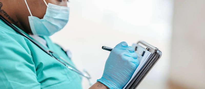 A pediatric clinician in scrubs taking notes during an in-person consultation.